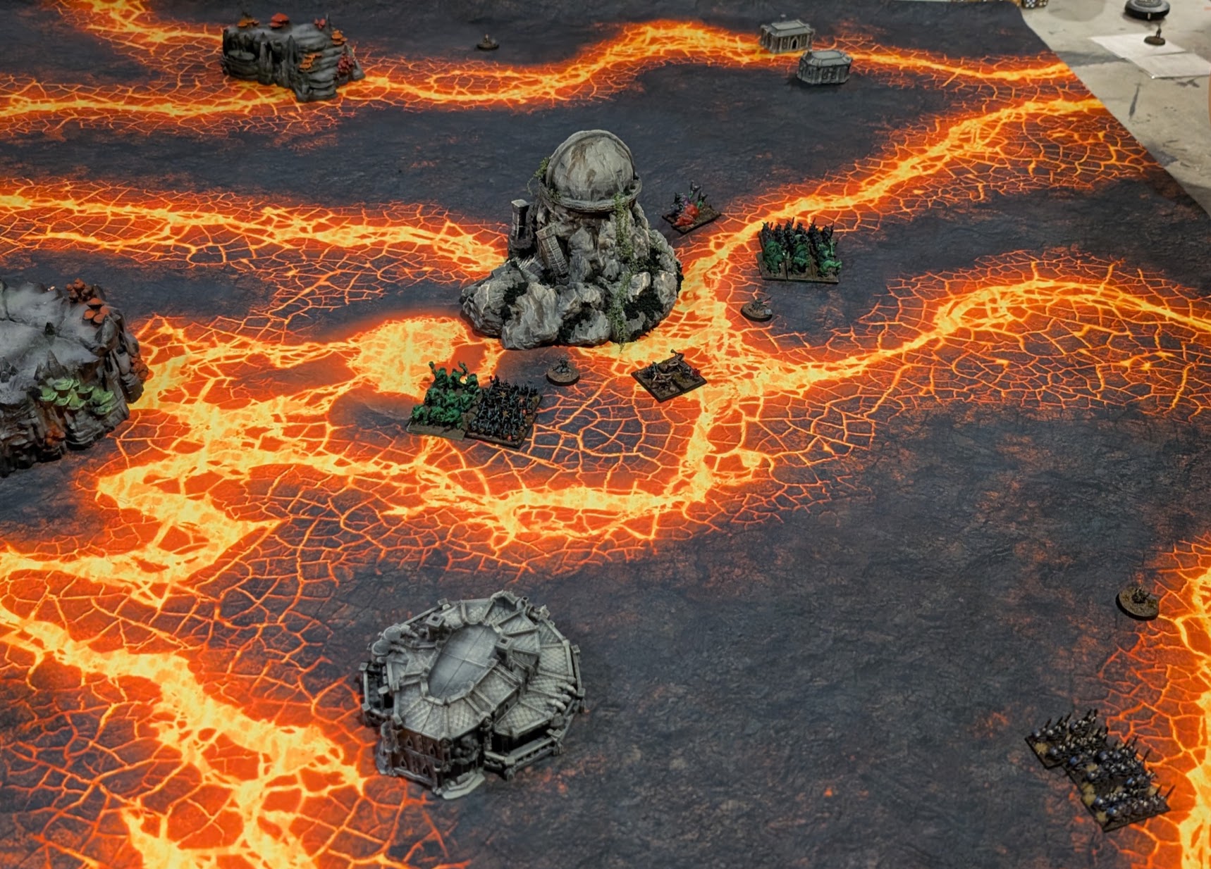 Warmaster units spread across a lava-patterned battlefield dotted with rocky outcrops and dwarf statues.